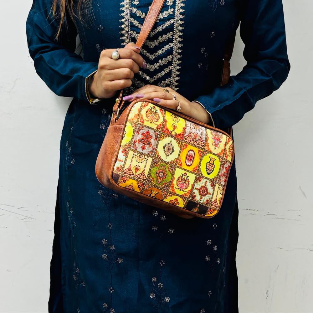 The person is holding a vibrant handbag with Mughal-style mosaic print and a brown strap. They are wearing a dark blue, embroidered dress adorned with rings on their fingers, standing against a plain white wall.