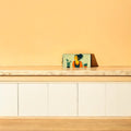 A vibrant yellow wall featuring a colorful painting, with a stylish purse placed nearby, adding a touch of elegance.